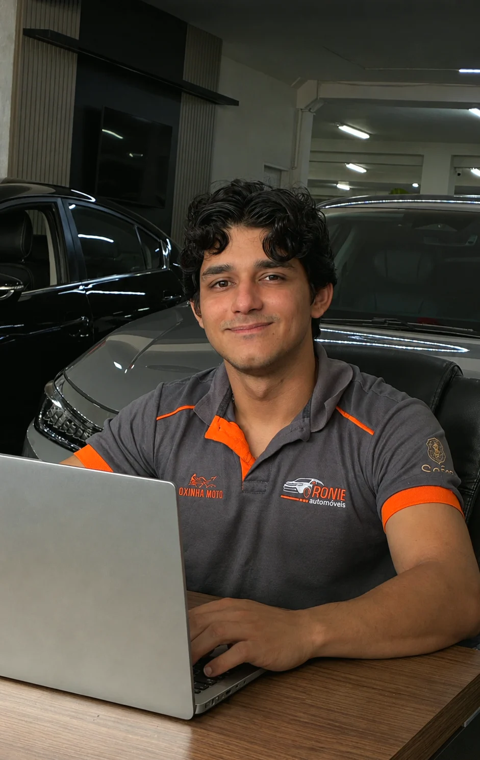 Neto, com o uniforme da Ronie Automóveis, no notebook da loja, com carros atrás